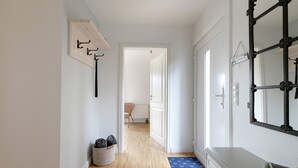 Un pasillo con piso de madera, una pared blanca con perchero, una cesta con objetos, un tapete azul y un aparador con espejo.