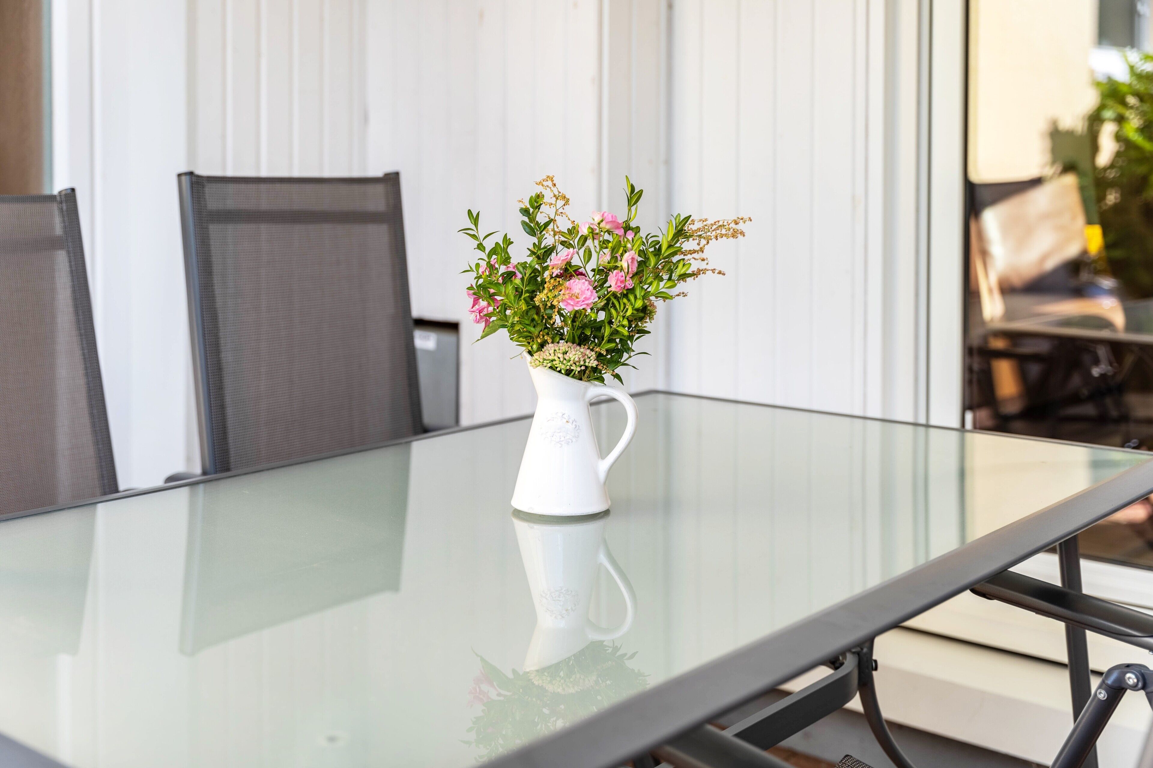 Une table dont le plateau est en verre, sur laquelle est posé un vase blanc contenant des fleurs roses.
