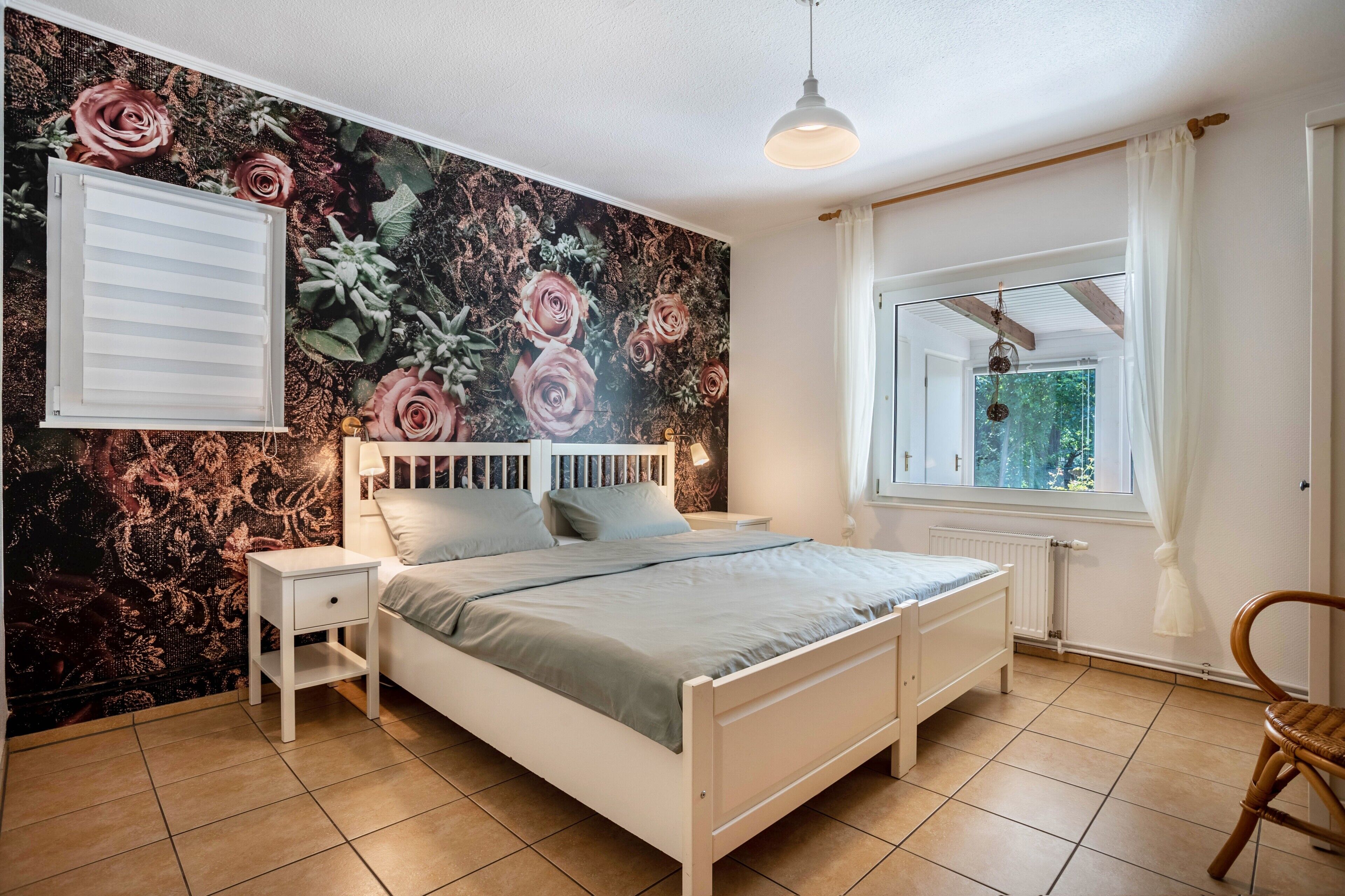 A bedroom with a bed, bedside tables, a window with curtains, a radiator, and a wall with a floral mural.