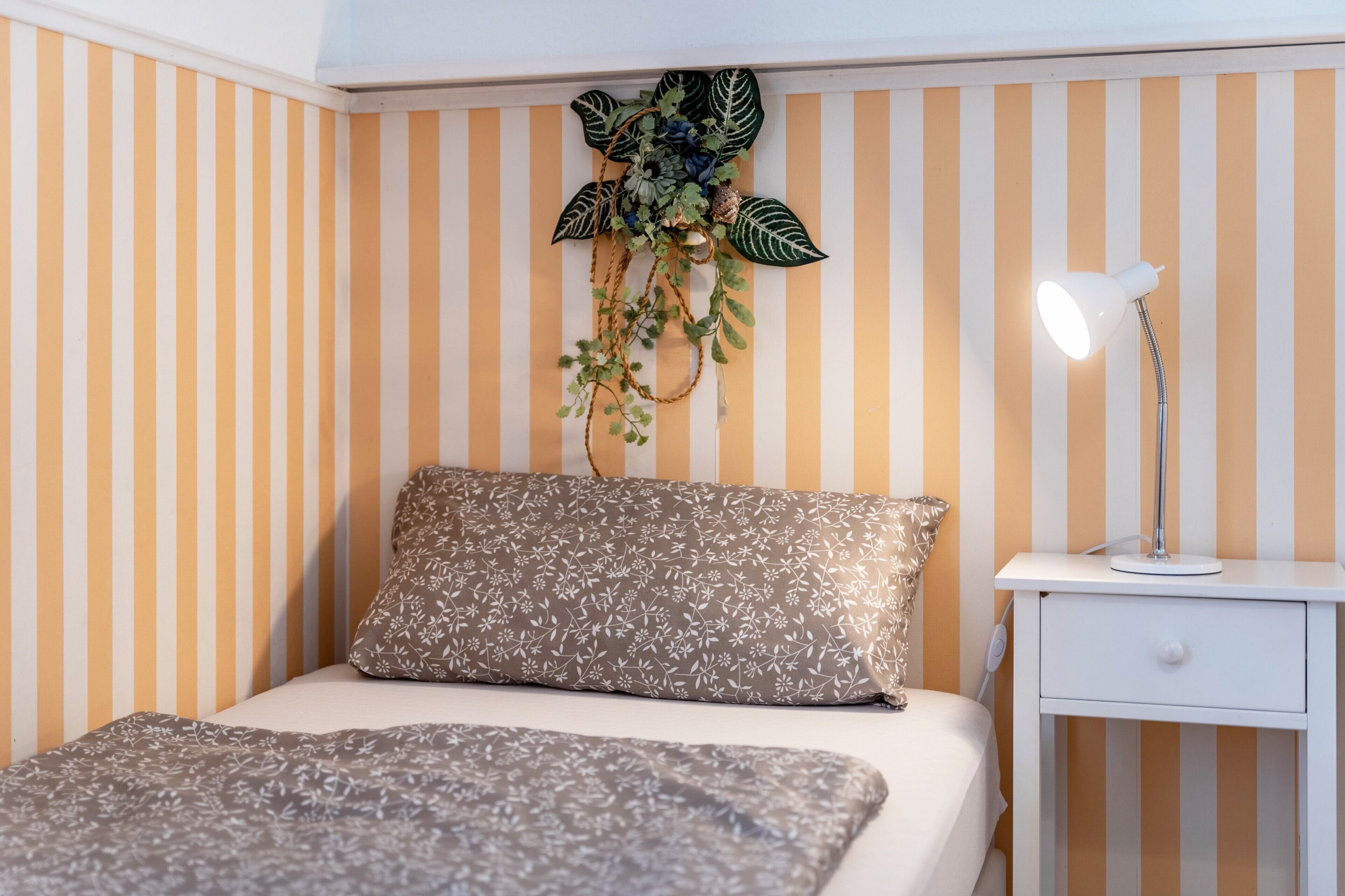Ein Bett mit einem blumigen Kopfkissen und einer Decke, eine Nachttischlampe, ein kleiner weißer Nachttisch und eine Wandpflanze mit Blättern und Blüten.