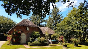A small brick house with a thatched roof, surrounded by well-maintained grass and flowering plants.