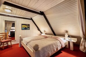 A bedroom with a sloped ceiling, a bed with a patterned blanket, a bedside table with a lamp, a chair, and a painting on the wall. - Holiday Apartment 'Rosenwohnung' with Shared Garden and Wi-Fi (Rodenäs)