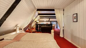 An attic bedroom with a sloped ceiling, a bed with a patterned bedspread, a chair, a lamp, and a framed picture on the wall.