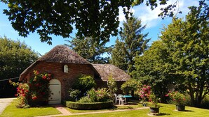 Ein kleines, aus Ziegeln gebautes Haus mit einem Strohdach, umgeben von gepflegtem Rasen und blühenden Pflanzen.