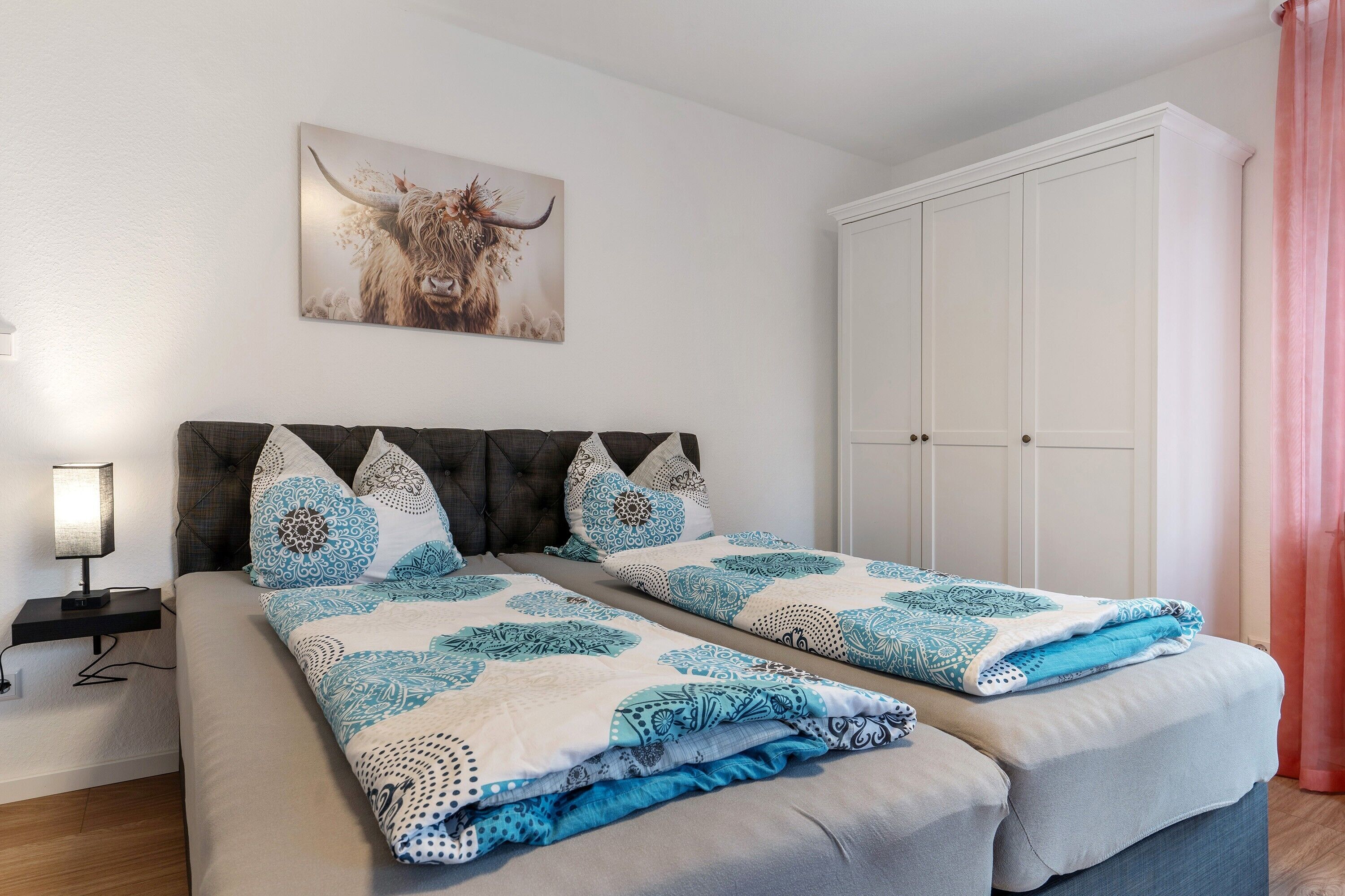 A double bed with patterned bedding, a headboard, a nightstand with a lamp, a wardrobe, and a bull painting on the wall.