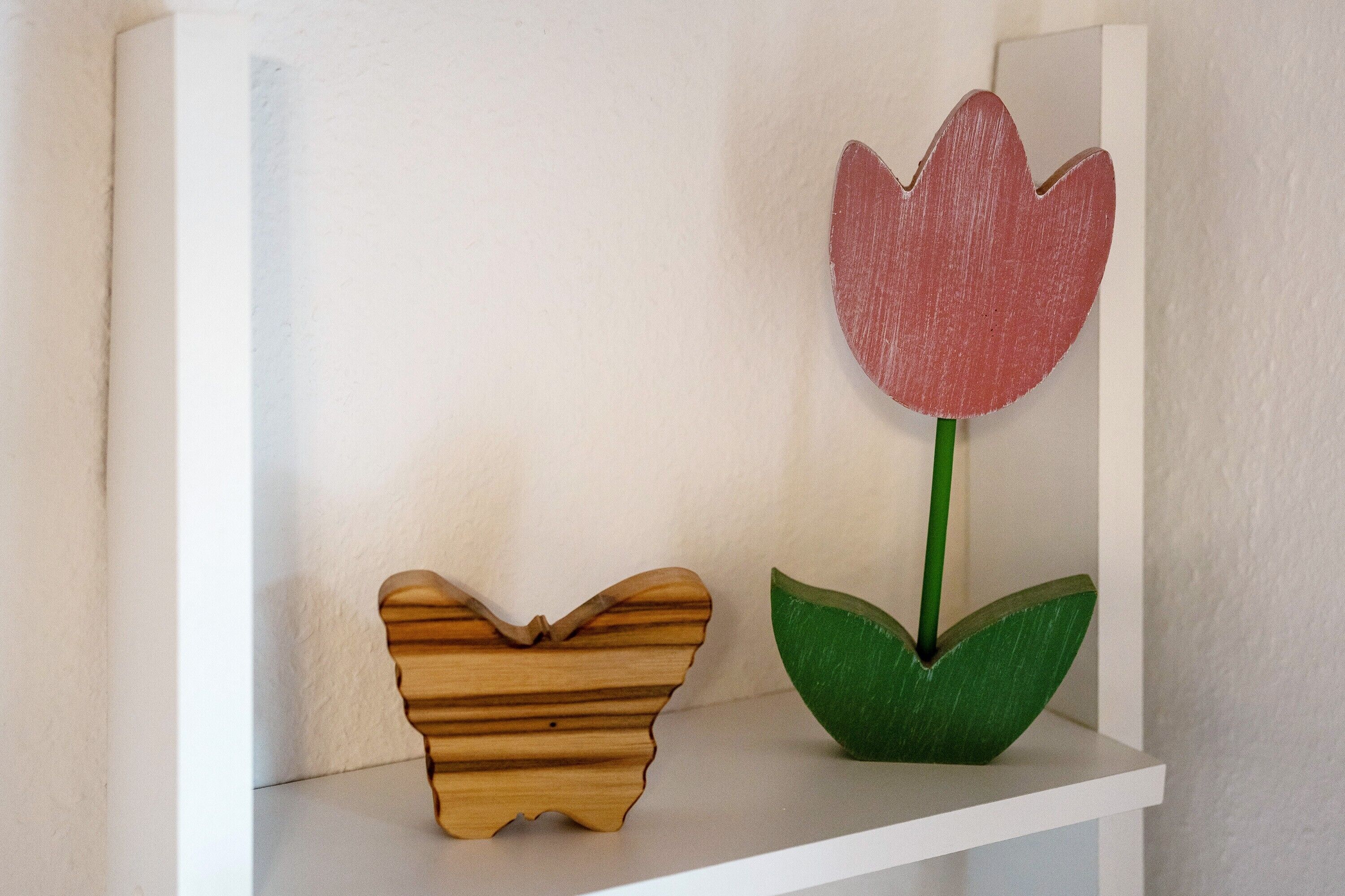 Un estante blanco con un objeto con forma de mariposa de madera y una flor pintada con tallo y hojas verdes.