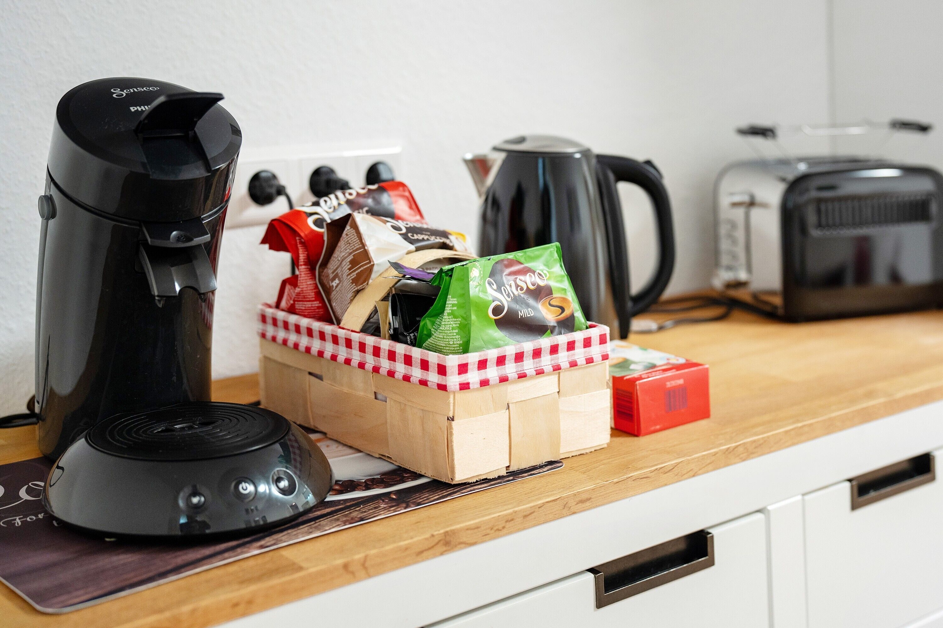 Un comptoir de cuisine avec une machine à café, une bouilloire, un grille-pain et un panier contenant des capsules de café et une boîte rouge.