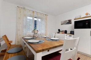 Un coin repas avec une table en bois dressée pour quatre personnes, des chaises blanches, et un espace cuisine équipé d’une machine à café et d’un four à micro-ondes.