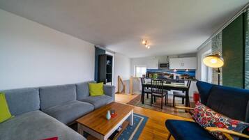 Una sala de estar moderna con un sofá, una mesa de centro y un sillón azul. Se observa un comedor con una mesa y sillas al fondo.