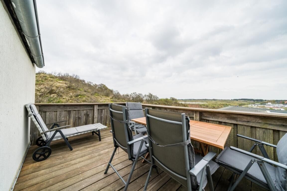 Una terraza de madera con mobiliario exterior, incluyendo sillas y una mesa, con vista a un paisaje con colinas y un edificio distante.