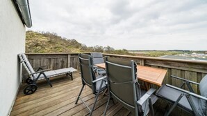 Una terraza de madera con mobiliario exterior, incluyendo sillas y una mesa, con vista a un paisaje con colinas y un edificio distante.