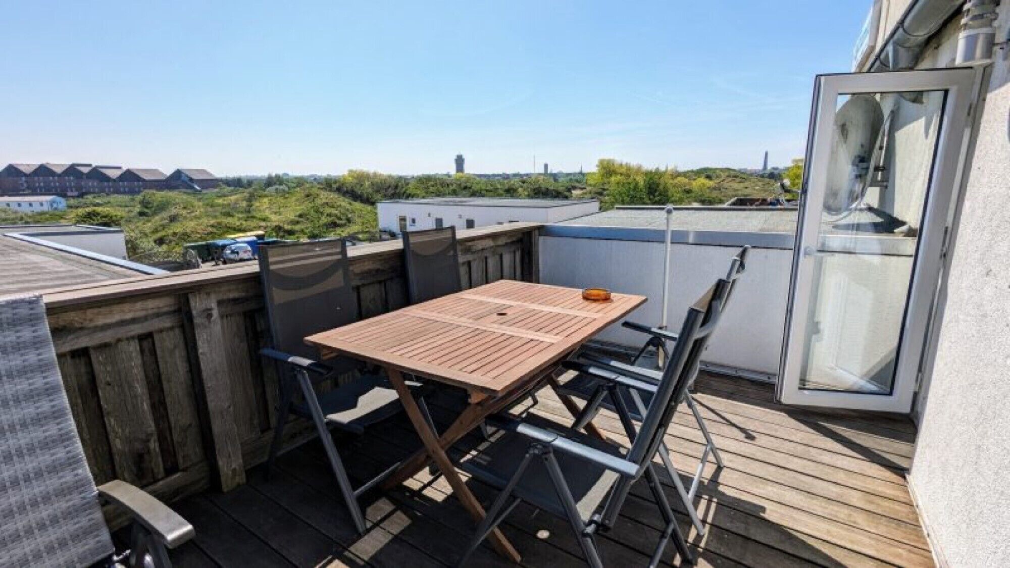 Une terrasse sur le toit avec une table et des chaises en bois, une porte vitrée et une vue sur les bâtiments et la verdure.
