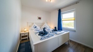 Un dormitorio con cama, mesita de noche, lámpara, ventana con cortinas y un pequeño cuadro en la pared.