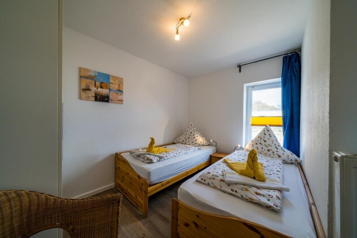 Un alojamiento pequeño de una sola habitación con dos camas de madera, una silla de mimbre y una ventana con cortinas.