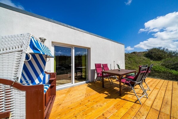 A modern building with a wooden deck, outdoor seating, and a large sliding glass door.