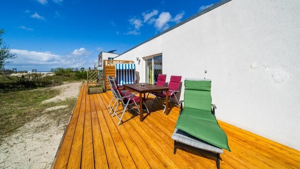 A wooden deck with a table and chairs, a green sunshade, and a white building.