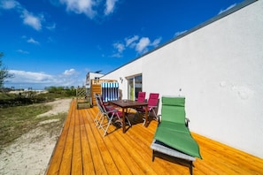 A wooden deck with a table and chairs, a green sunshade, and a white building.