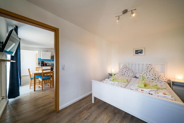 A bedroom with a white bed, a small bedside table, a wall-mounted light, and a door leading to a dining area.