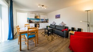 Eine moderne Wohnung mit Essbereich, einem Wohnzimmer inklusive Sofa und einem roten Sessel, einer dahinterliegenden Küche und einem großen Fenster mit Vorhängen.
