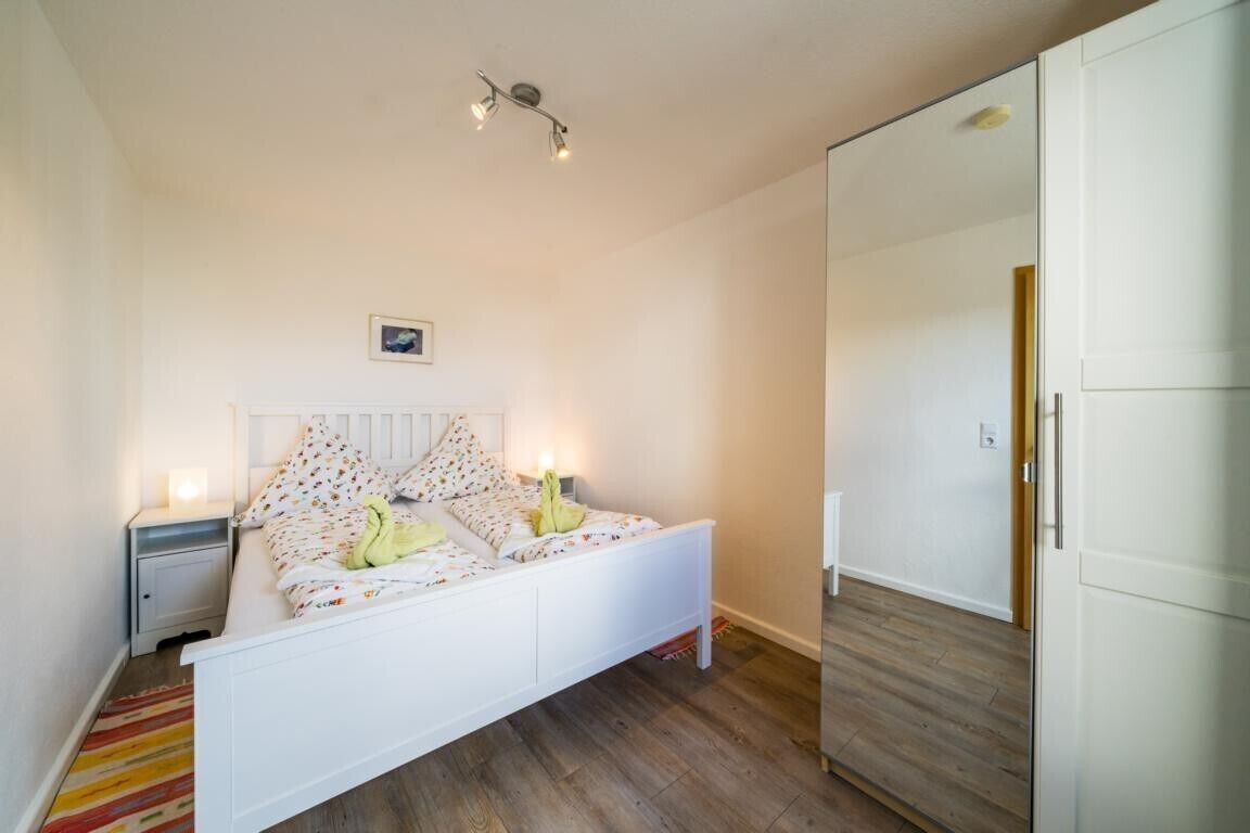 Un dormitorio con una cama blanca, una mesita de noche, una lámpara de pie, un pequeño cuadro en la pared y suelo de madera.