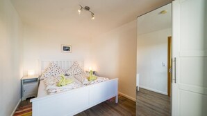 Ein Schlafzimmer mit einem weißen Bett, einem Nachttisch, einer Nachttischlampe, einem kleinen Bild an der Wand und einem Holzboden.