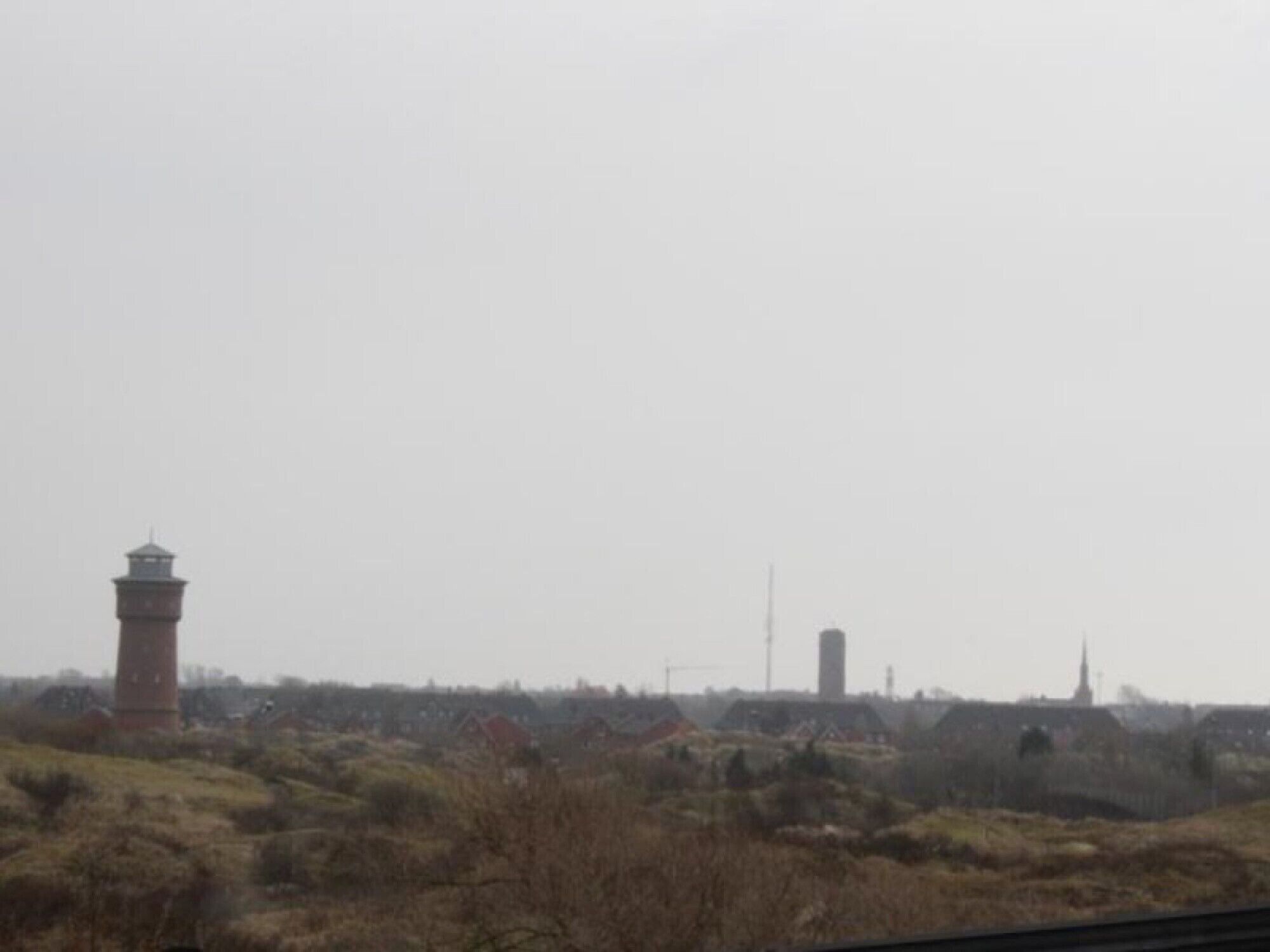 Un phare situé dans un paysage rural, avec une végétation clairsemée et quelques bâtiments en arrière-plan.