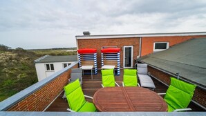 Een dakterras met een houten tafel en groene stoelen, een bakstenen gebouw en uitzicht op het landschap.