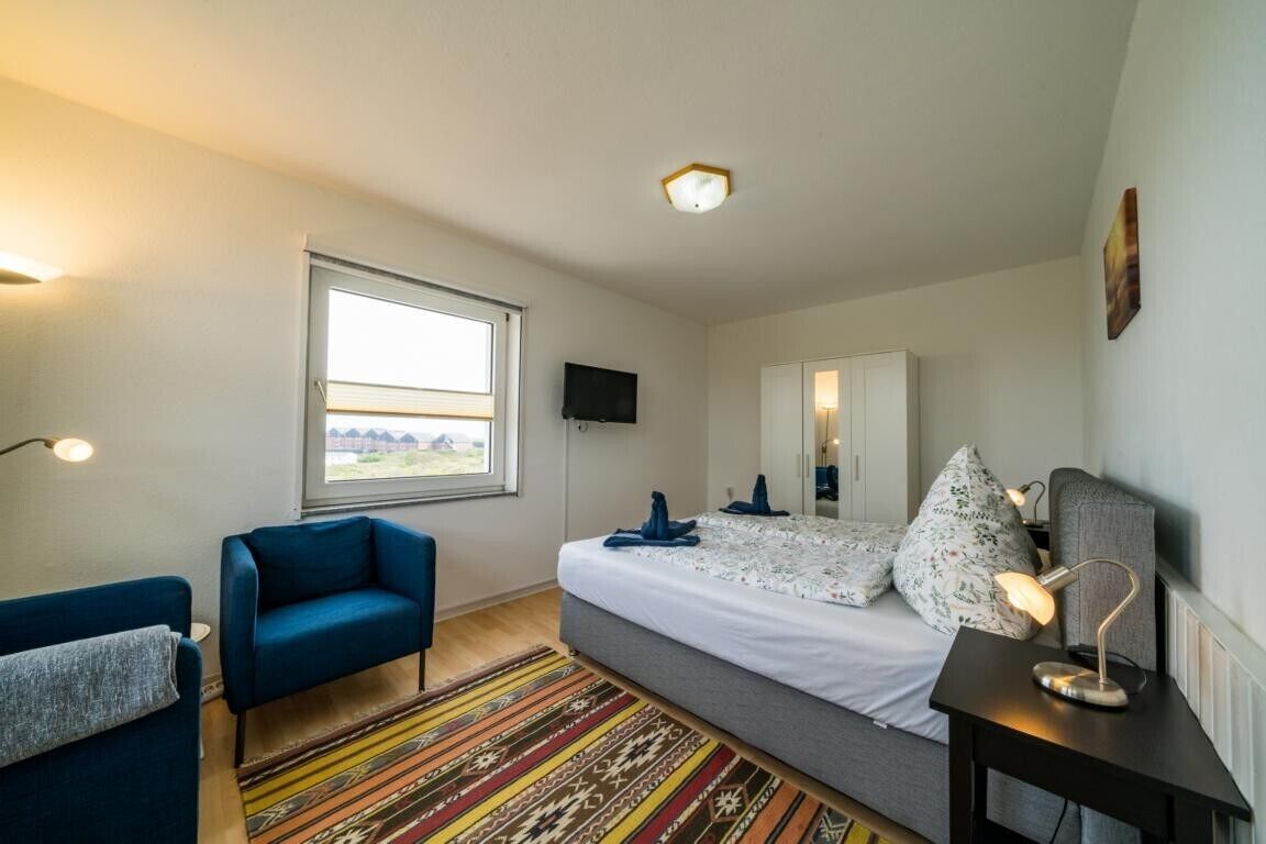 A bedroom with a bed, a blue chair, a bedside table with a lamp, a window, and a television on the wall.