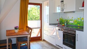 A kitchen with white cabinets, a wooden table with chairs, a window with yellow curtains, and a view of greenery outside. - Apartment 'Olde Toornblick' with Balcony and Wi-Fi (Borkum)