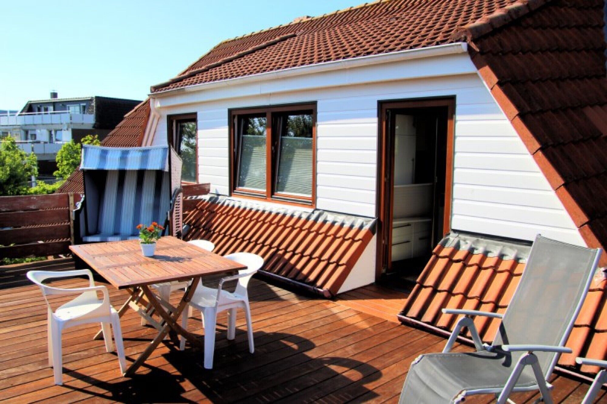 Une terrasse en bois avec une table et des chaises, un petit jardin et un bâtiment doté d’un toit en tuiles.