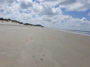 A wide sandy beach with footprints leading to the water, a clear sky, and dunes in the background.