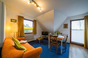 Una habitación con sofá, una mesa y sillas, televisor y ventana con cortinas.