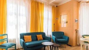 Um quarto com um sofá azul, duas cadeiras iguais, uma mesa redonda pequena e cortinas amarelas.