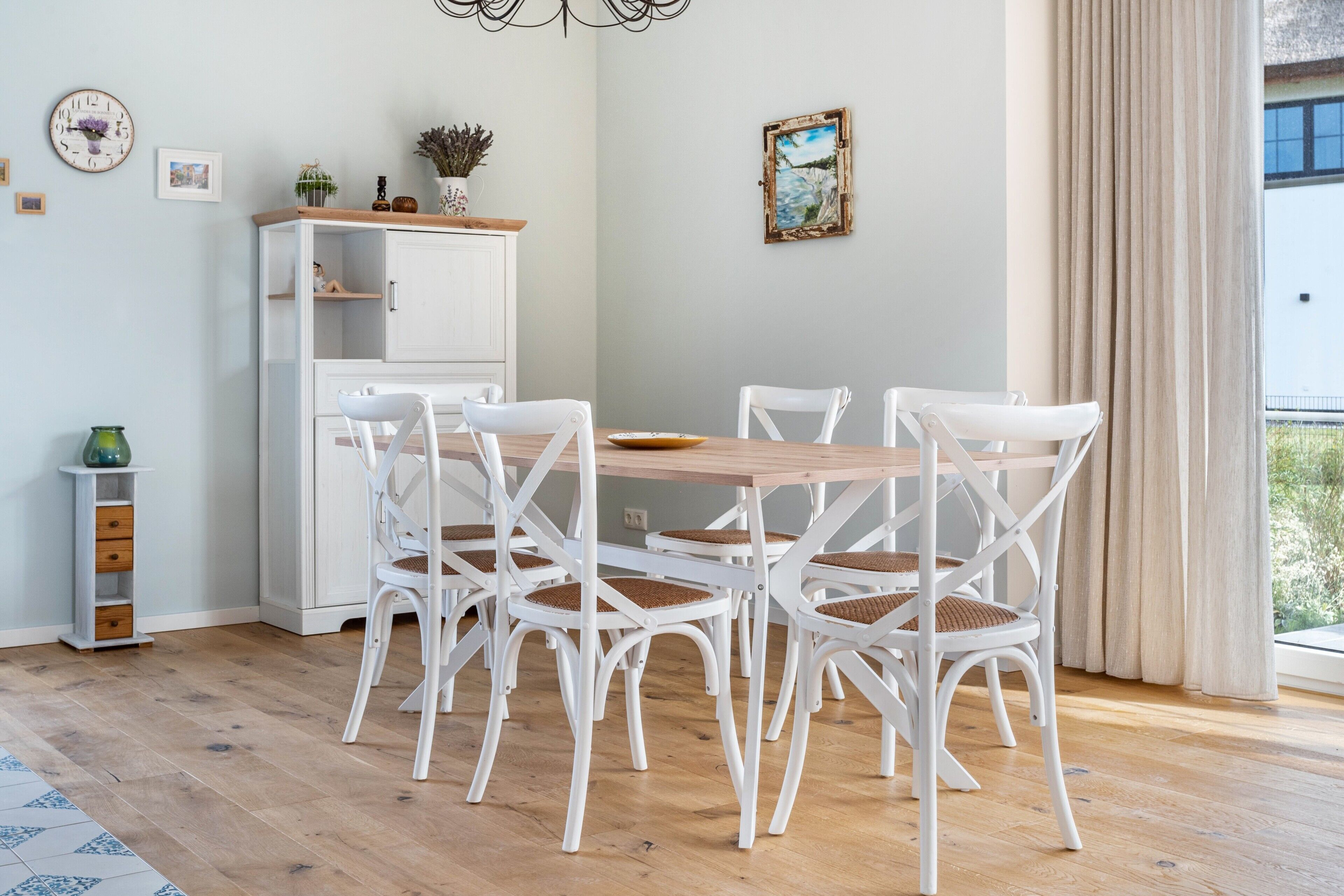 A dining area with a wooden table and white chairs, a white cabinet with shelves, a clock, and a painting on the wall.