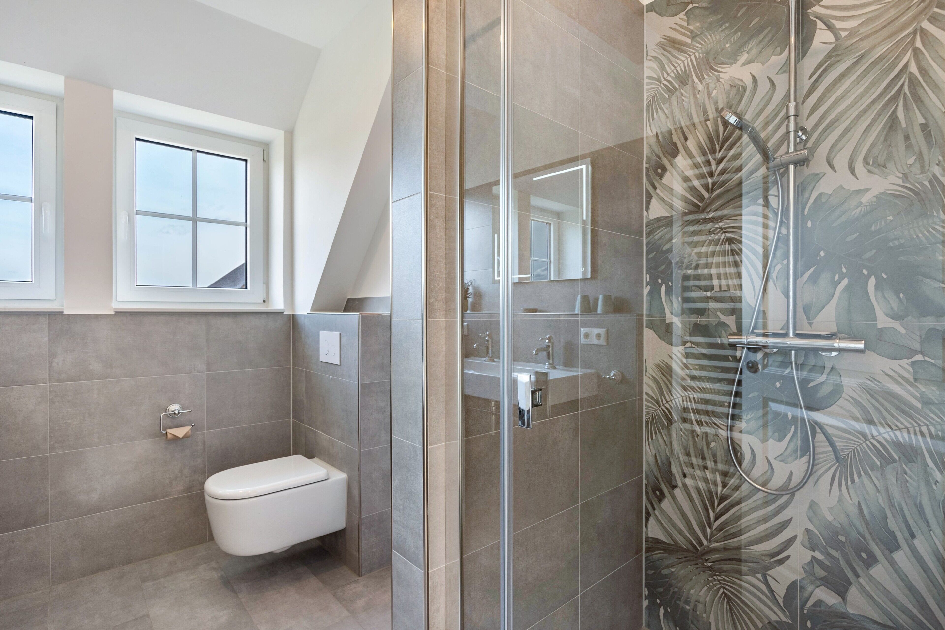 Ein modernes Badezimmer mit Toilette, einer in Glas abgeschirmten Dusche mit Regendusche und einem Waschbecken mit Spiegel.
