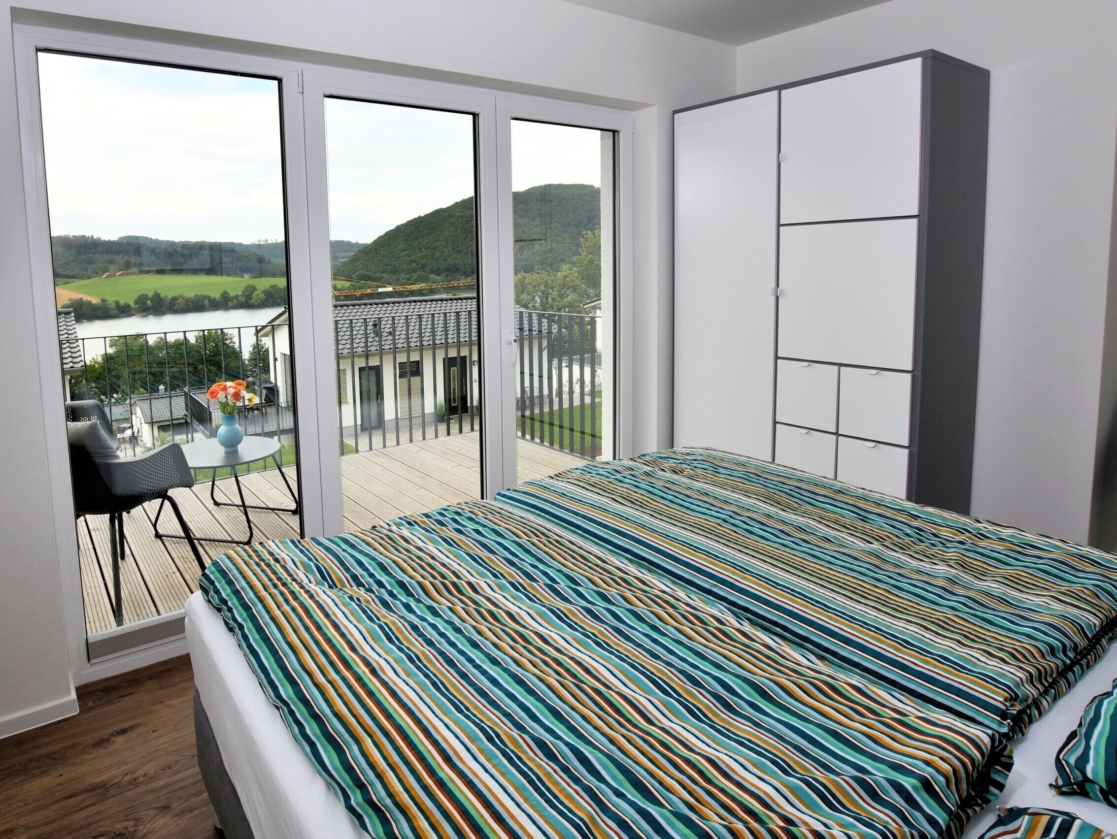Een slaapkamer met een groot bed, een balkon met uitzicht op een meer en heuvels, een kleine tafel met stoelen en een kledingkast.