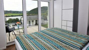 Een slaapkamer met een groot bed, een balkon met uitzicht op een meer en heuvels, een kleine tafel met stoelen en een kledingkast.