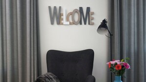 A modern room with a grey upholstered chair, a matching ottoman, a teal side table with a vase of flowers, and a 'WELCOME' sign on the wall. - Holiday Home 'Christa' with Lake View, Private Terrace and Wi-Fi (Diemelsee)