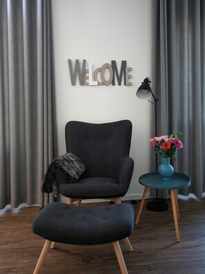 A modern room with a grey upholstered chair, a matching ottoman, a teal side table with a vase of flowers, and a 'WELCOME' sign on the wall.