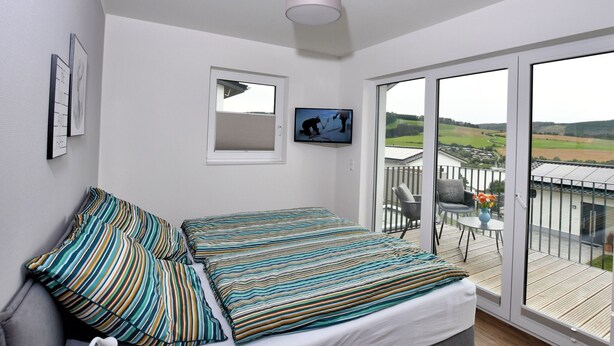 A bedroom with a bed, pillows, a TV, a window, and a balcony with a table and chairs. - Holiday Home 'Christa' with Lake View, Private Terrace and Wi-Fi (Diemelsee)