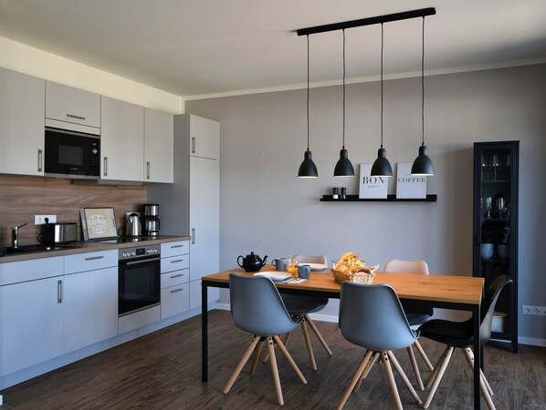 A modern kitchen with white cabinets, a microwave, and a coffee maker. There is a dining table with chairs and a hanging light fixture above.