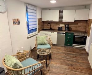 A compact kitchen with white cabinets, a dishwasher, a coffee maker, and a built-in oven. There is a wicker chair with a cushion and a small round table. - Holiday Home 'Kleines Haus' with Private Terrace and Wi-Fi (Bernau bei Berlin)