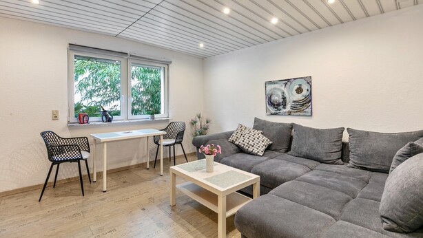 A modern living room with a grey sofa, a white coffee table, a dining table with chairs, and a window with a view of greenery.