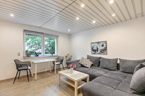 A modern living room with a grey sofa, a white coffee table, a dining table with chairs, and a window with a view of greenery. - Holiday Home 'Kleines Haus' with Private Terrace and Wi-Fi (Bernau bei Berlin)