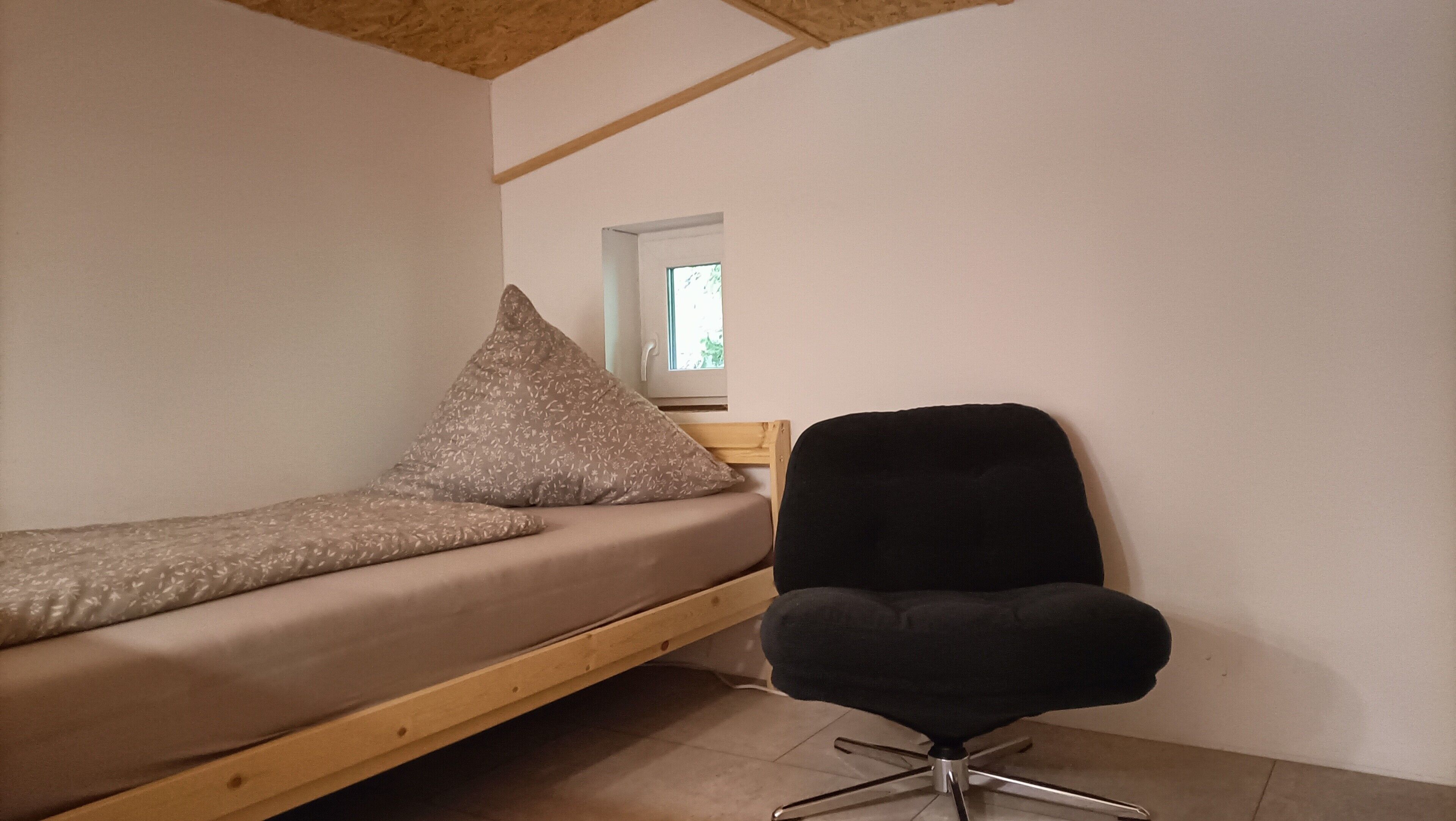Ein kleines Zimmer mit einem Einzelbett, einem Sessel und einem Fenster.