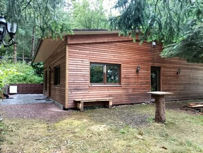 Une cabane en bois avec une fenêtre, une porte et un petit espace extérieur aménagé pour s’asseoir.