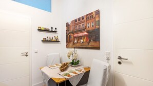 A small room with a dining table, chairs, a painting of a street scene, and shelves with bottles.
