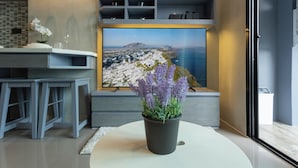 Uma sala de estar moderna com uma televisão de tela plana grande fixada na parede, um vaso de lavanda sobre uma mesa branca, e um design minimalista com tons de cinza e um tapete branco.