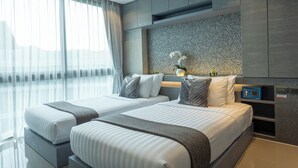 Quarto de hotel moderno com duas camas, uma janela grande com cortinas translúcidas, um painel decorativo na parede e uma mesa de cabeceira com luminária.
