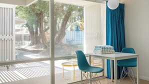 A room with a large window, a table with chairs, and a view of trees and a blue fence outside. - Patio, Pool, Firepit | Queen Studio at Monkey Tree (Palm Springs)